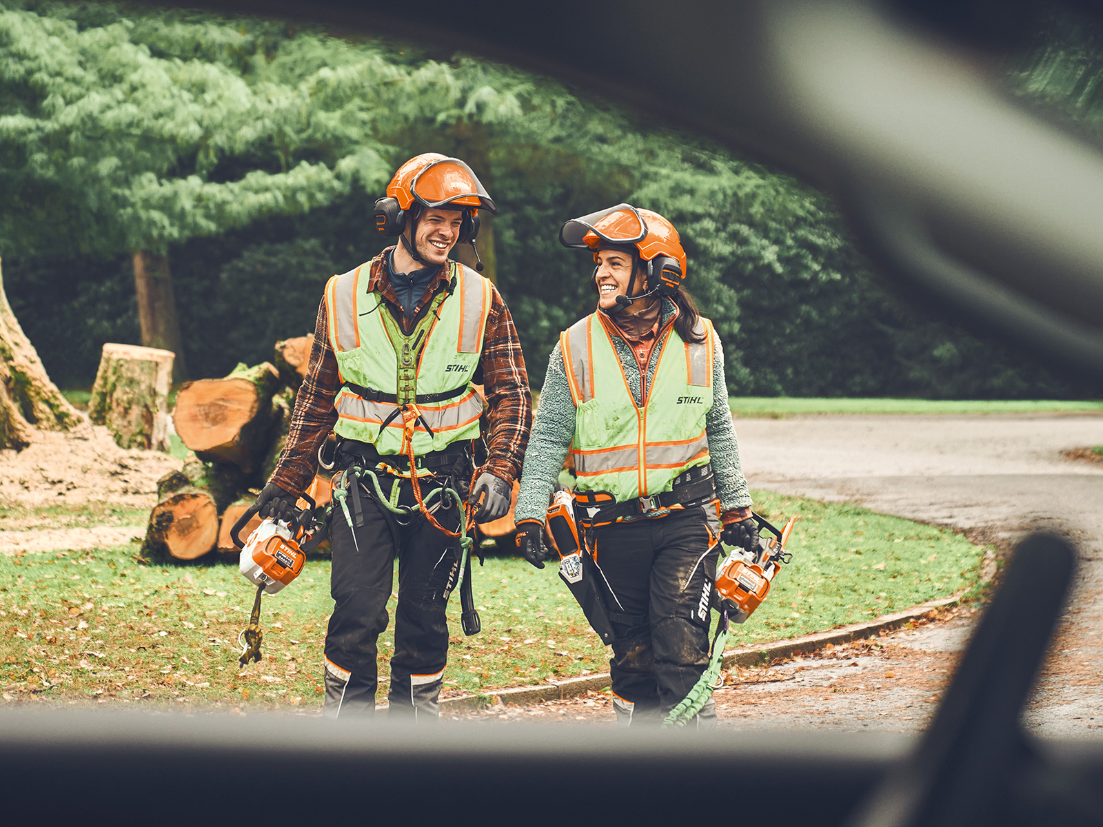 Ein Mann und eine Frau mit STIHL Akku-Motorsägen und einem STIHL Akku-Gehölzschneider laufen lächelnd in Richtung eines Autos  